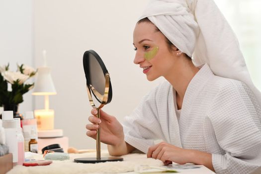 Beautiful caucasian woman applying anti fatigue under eyes and looking at mirror on dresser table. Beauty treatment concept.