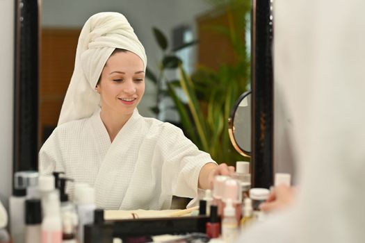 Millennial caucasian woman in bathrobe sitting front of mirror and applying cream, making daily beauty routine at home.