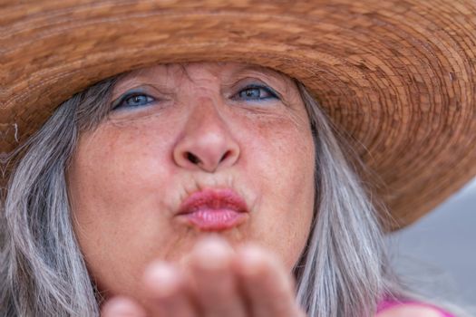 close-up of an older woman with a hat, white hair and blue eyes blowing a kiss with her hand