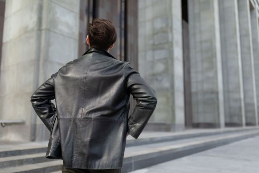 a mature man in a leather jacket is standing looking at the buildings in the city, the view from the back.