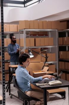 Woman supervisor looking at cargo stock on laptop computer while checking fashion blouse status before start preparing clients packages. Employee working at warehouse delivery department