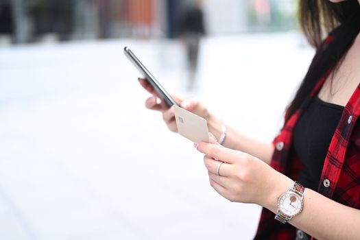 Close up view female hand holding credit card and using smart phone while sitting at outdoor.