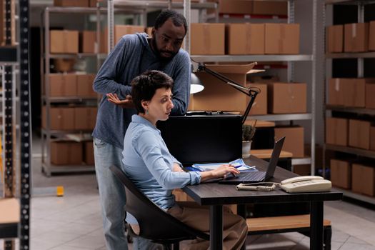 Warehouse workers analyzing checklist on laptop computer searching for metallic box order before shipping package. Multi ethnic employees analyzing at delivery logistics while working in storehouse