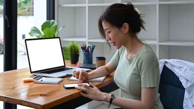 Young woman sitting in front of laptop computer and using smart phone.