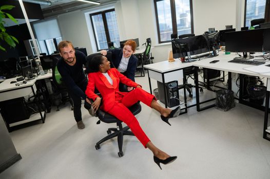 Caucasian red-haired woman, bearded caucasian man rolled African American young woman on office chair. Colleagues have fun at work