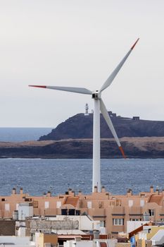 Wind turbine located in small town near blue rippling sea against lighthouse on hill