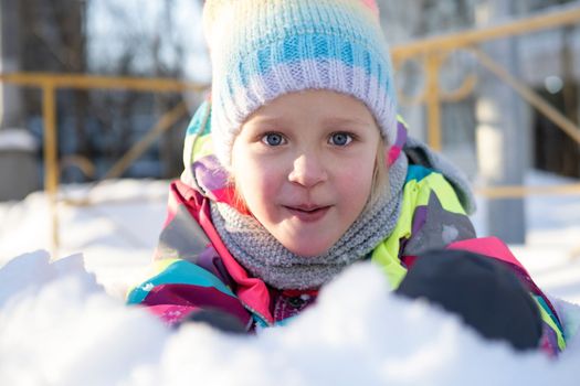 Portrait of cute smiling girl in colorful knitted hat and scarf lying on snow and looking at camera