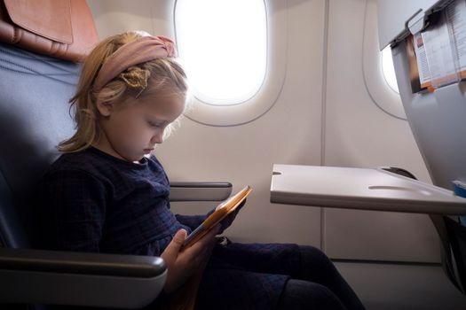Side view of focused preteen girl in dress sitting on seat in plane and watching cartoon via tablet