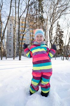 Cheerful girl in bright colorful outerwear and hat standing on snowy road in yard near residential building