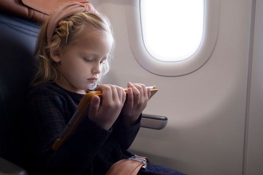 Concentrated preteen girl with blond hair wearing headband sitting on passenger seat in plane and watching cartoon via tablet