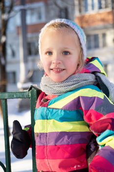 Adorable positive preteen girl in colorful outerwear and knitted hat standing near metal fence on playground against residential multistory building during winter time