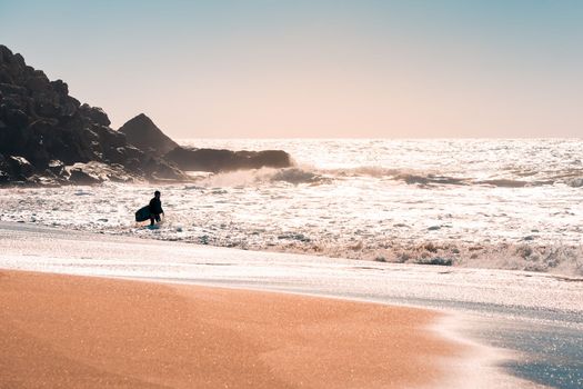 silhouette of a man with his board at the seashore preparing to surf, the sunlight reflects on the water and the sand, at the background you see the rocks of a cliff, the sky is clean and clear