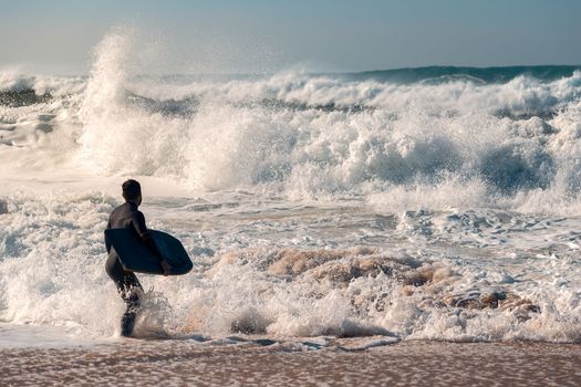 Man on the seashore prepares to surf, wears a black neoprene wetsuit and in his hand has a bodyboard, water covers him to the knee while defies the rough sea breaking powerful