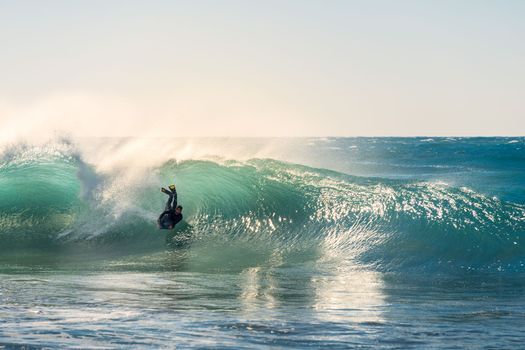 Man surfing rides the barrel of a big wave that breaks with energy and power, sunlight reflects golden on the turquoise surface of the sea and in the huge pile of foam, wind rises columns of water