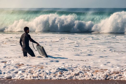 Man on the seashore prepares to surf, wears a black neoprene wetsuit and in his hand has a bodyboard, water covers him to the knee while watching a huge wave breaking powerful, seawater is turquoise