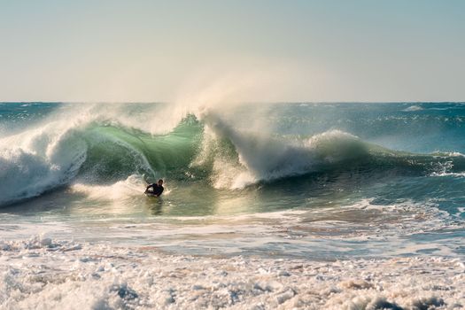 man surfing rides a big wave that breaks with a lot of energy and power, sunlight reflects golden on the turquoise surface of the sea and in the huge pile of foam, wind rises plumes of water