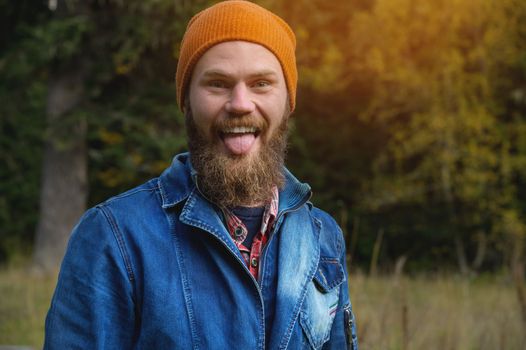 An attractive bearded man in a red hat and a denim jacket grimaces showing his tongue at the camera in the forest. funny portrait.