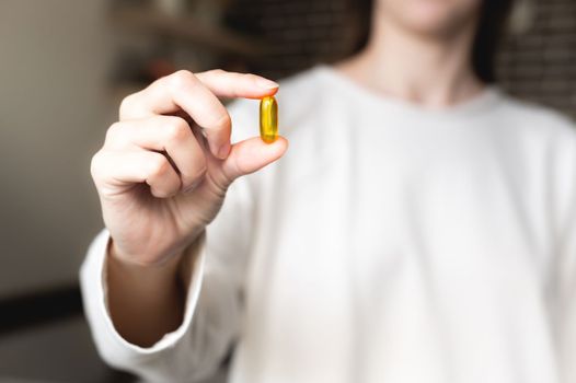 A woman's hand holds an Omega-3 capsule against the background of herself in a white sweater, the background is in blur. Useful fish oil capsule tablets.