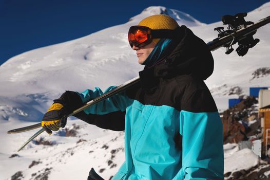 A portrait of a professional skier in an orange ski goggles stands with skis on his shoulder against the backdrop of snow-capped mountains. ski resort.