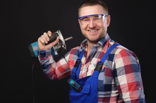 A smiling man in a protective suit and a plaid shirt wearing eye protection goggles stands and holds a repair tool in his hands. Portrait on a black background.