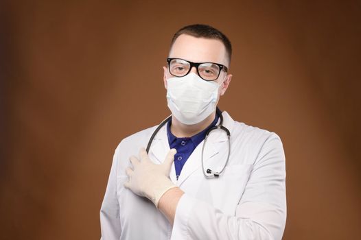 A doctor in a medical mask, with a stethoscope around his neck and wearing gloves, stands in a white coat and holds his hand on his heart. Portrait of a doctor.