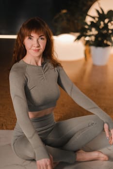 A woman in a gray tracksuit sits on a yoga mat in a lotus position in the gym.