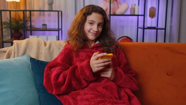Preteen girl texting share messages content on smartphone social media applications online watching relax movie, news. Young child, kid at home in night evening living play room sitting on orange sofa