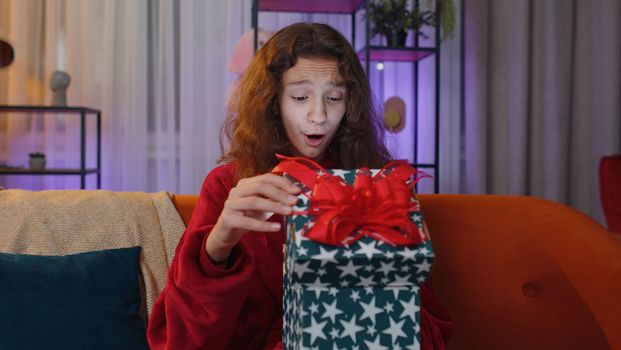 Preteen girl opening gift box with light glow inside, smiling joyfully, looking amazed satisfied with nice present, unexpected birthday surprise. Young child kid at home in evening living play room