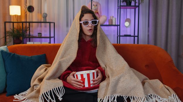 Excited preteen girl in 3D glasses eating popcorn and watching interesting tv serial, sport game, film, online social media movie content. Young child kid at home in evening living play room on couch