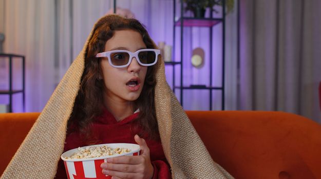 Excited preteen girl in 3D glasses eating popcorn and watching interesting tv serial, sport game, film, online social media movie content. Young child kid at home in evening living play room on couch