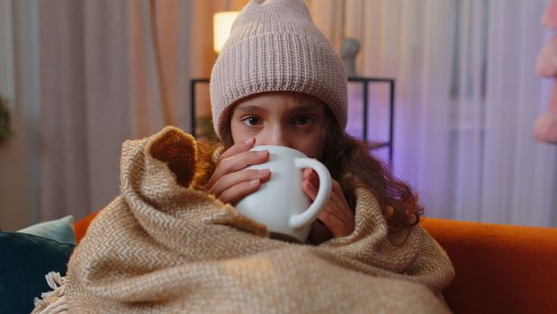 Sick preteen girl wear hat wrapped in plaid sit alone shivering from cold on sofa drinking hot tea in unheated apartment without heating due debt. Unhealthy child feeling discomfort try to warming up