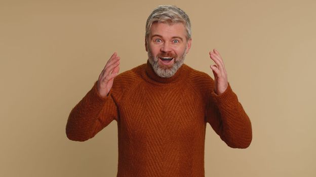 Oh my God, Wow. Excited amazed winner man raising hands in surprise looking at camera with big eyes, shocked by sudden victory. Sincere senior middle-aged guy isolated alone on beige studio background