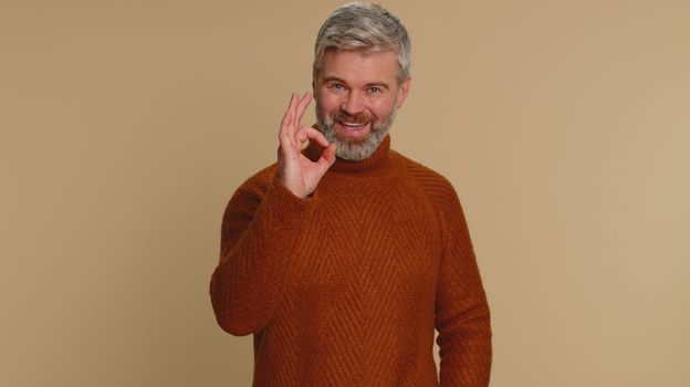 Ok. Joyful happy middle-aged man in sweater looking approvingly at camera showing approve feedback gesture like sign positive something good. Senior guy on beige background. People sincere emotion
