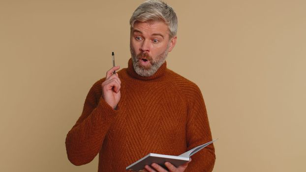 Thoughtful journalist middle-aged old man making notes, writing down thoughts with pen into notepad notebook diary, to do list, good idea. Senior mature guy isolated alone on beige studio background