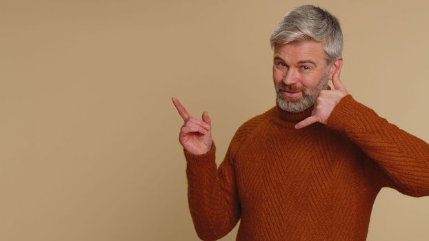 Call us, here is contact number. Senior middle-aged man in sweater looking at camera doing phone gesture like says hey you call me back, conversation. Caucasian mature guy isolated on beige background