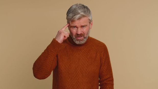 You are crazy, out of mind. Middle-aged elderly old man pointing at camera and showing stupid gesture, blaming some idiot for insane plan. Senior mature guy isolated alone on beige studio background