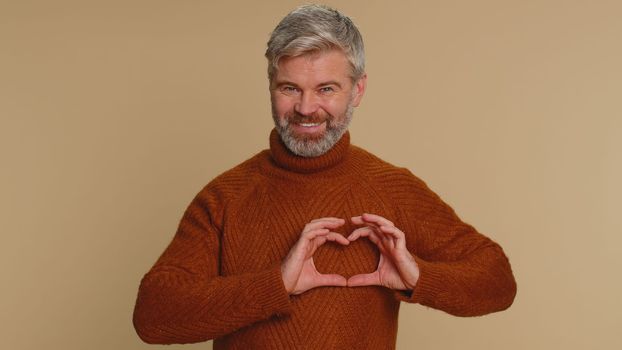 Man in love. Smiling middle-aged man 45 years old makes heart gesture demonstrates love sign expresses good feelings and sympathy. Senior mature adult guy isolated alone on beige studio background
