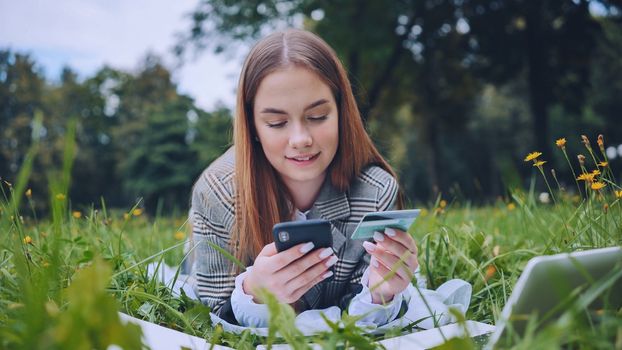 A young girl makes a purchase with her phone and bank card while lying on the grass in the park