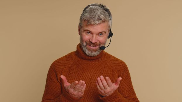 Smiling middle-aged man wearing headset, freelance worker, call center or support service operator helpline, having talk with client or colleague, communication support. Senior guy on beige background