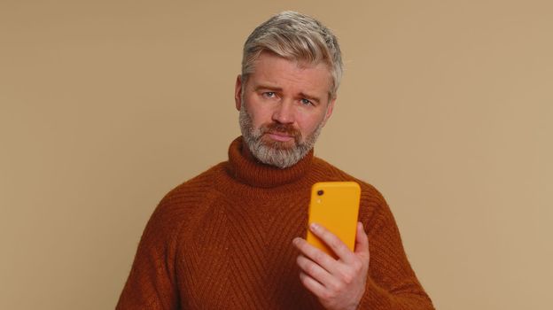 Middle-aged old man use mobile cell phone typing browsing, loses becoming surprised sudden lottery results, bad fortune, loss, fail. Senior mature guy isolated alone on beige studio wall background