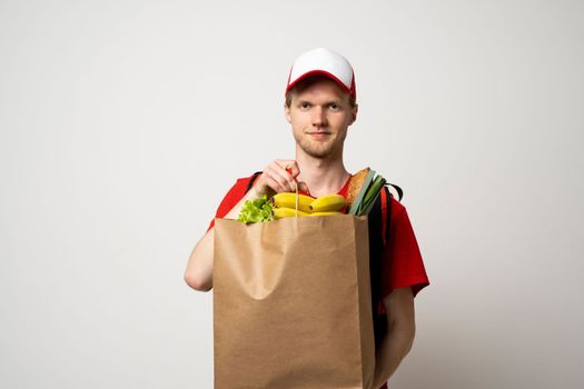 Handsome delivery man in a cap, courier carrying package bag of grocery food and drinks from store, supermarket isolated on white studio background. Food delivery service concept