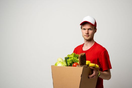 Side view delivery man in red uniform delivering food, groceries, vegetables, drinks in a paper box to a client at home. Online grocery shopping service concept