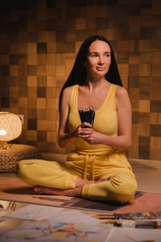 A girl in yellow clothes sitting on the floor holding pencils for drawing.
