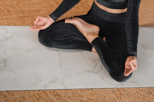 Close-up of the legs and arms of a woman in a black tracksuit sitting on a yoga mat in a lotus position in the gym.