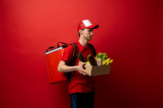 Side view delivery man in red uniform on a red background delivering food, groceries, vegetables, drinks in a paper box to a client at home. Online grocery shopping service concept