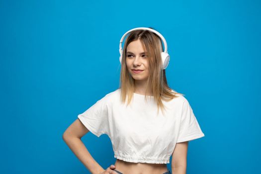 Beautiful attractive young blond woman wearing white t-shirt and glasses in white headphones listening music and smiling on blue background in studio. Relaxing and enjoying. Lifestyle