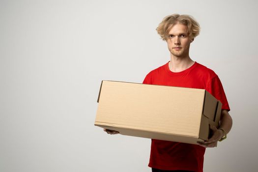 Portrait of a young delivery man, courier in red cap and t-shirt standing with parcel post box isolated over white background
