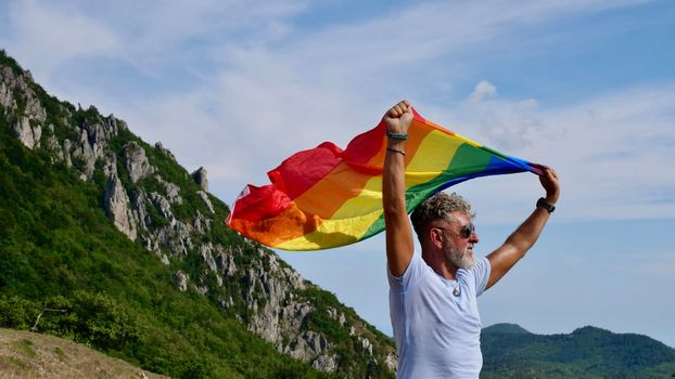 Portrait of a gray-haired senior elderly Caucasian man bisexuality with a beard and sunglasses holding a rainbow LGBTQIA flag on nature. Celebrates Pride Month, Rainbow Flag Day, gay parade