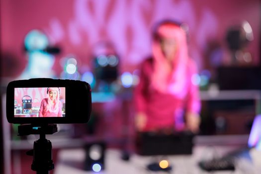 Artist with pink hair standing at table in night club performing techno song using mixer console while filming music session with professional camera. Dj posting performance video on her channel