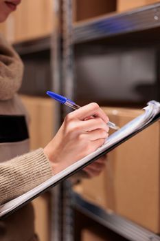 Post office warehouse employee searching parcel, checking postal form. Young caucasian woman supervising cardboard boxes in mail sorting center and writing on clipboard close up